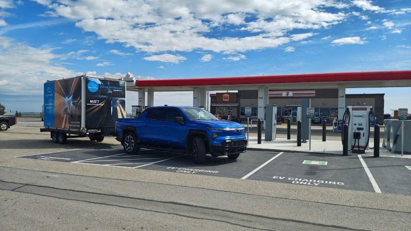 Blue electric truck with a trailer parked at an EV charging station near a convenience store.