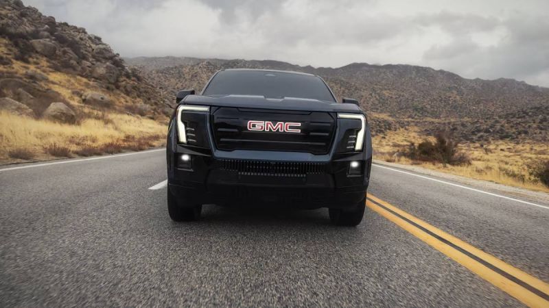 Black GMC pickup truck, front view on desert highway, featuring LED headlights, bold grille, rocky mountain landscape background.