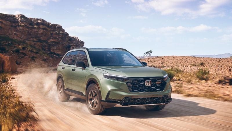 Green Honda CR-V SUV driving on dusty desert road with rocky cliff backdrop