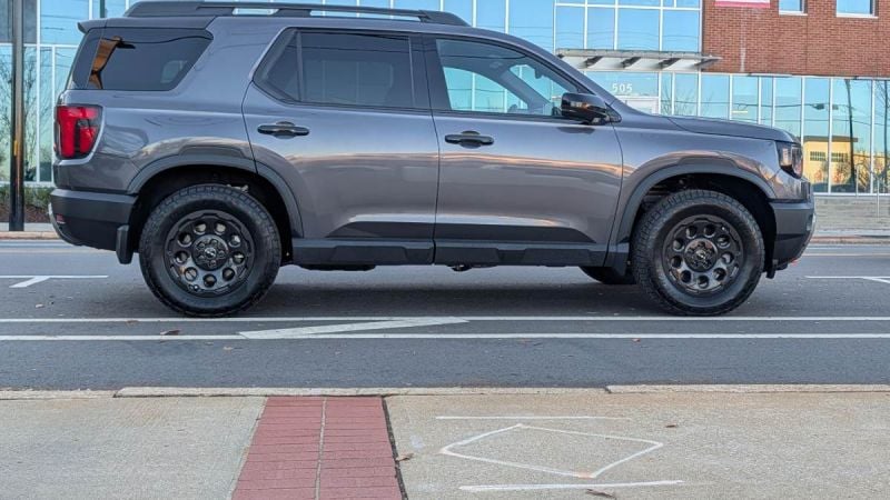 2026 Honda Passport TrailSport Elite in gray shown in full side profile parked on a city street with modern buildings in the background.