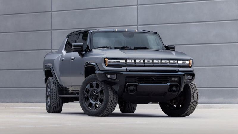A sleek, grey Hummer EV pickup truck parked in front of a modern, minimalist grey wall, showcasing its bold design and large tires.