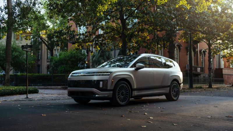 A silver Hyundai Ioniq 9, viewed from a front three-quarter angle, parked on a tree-lined historic city street with brick buildings in the background.