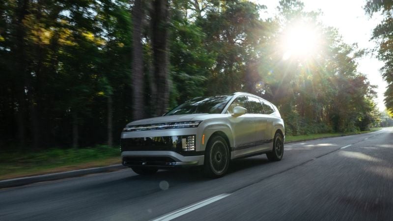 Silver SUV driving on tree-lined road with sunlight streaming through forest canopy