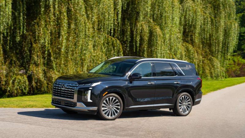2026 Hyundai Palisade in black, photographed at three-quarter front angle on paved road beneath weeping willow trees, featuring distinctive split LED headlights and large grille.