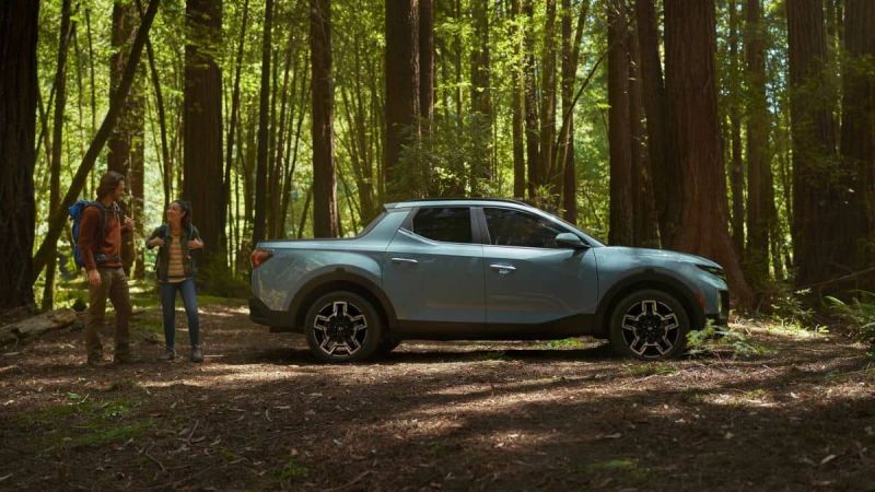 A light blue Hyundai Santa Cruz is parked in a sunlit forest. Two people with backpacks talk nearby, surrounded by tall trees, conveying a sense of adventure.