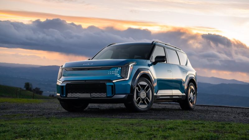 Blue Kia EV9 SUV parked on hillside against dramatic sunset sky with mountains.
