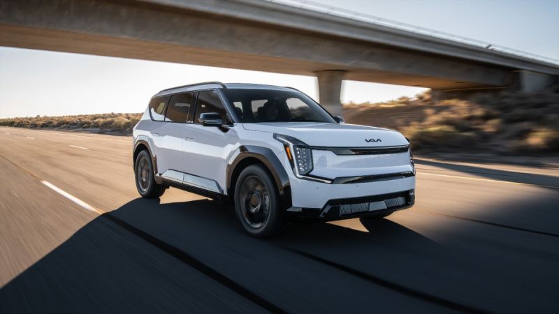 White 2026 Kia EV9 SUV driving under highway overpass, angular design with dynamic motion blur