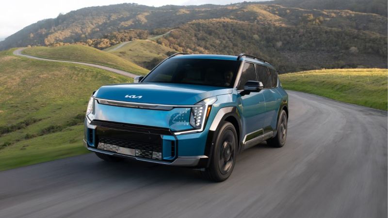 A blue 2026 Kia EV9 with modern styling drives on a winding road through green hills and mountains, conveying a sense of adventure and freedom.