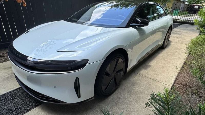 A sleek white electric car with black accents parked on a concrete driveway, surrounded by greenery and a dark wooden fence.
