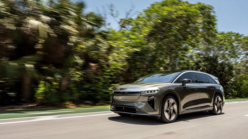 2026 Lucid Gravity shown in three-quarter front view, driving on tree-lined road with distinctive LED lighting and aerodynamic wheels.