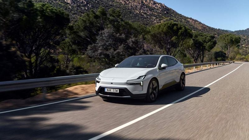 A sleek, white electric car drives along a winding road, surrounded by lush trees and a mountainous backdrop under clear skies.