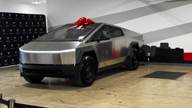 2026 Tesla Cybertruck RWD front three-quarter view indoors with a red bow on the roof, showcasing stainless steel body and angular electric pickup design.