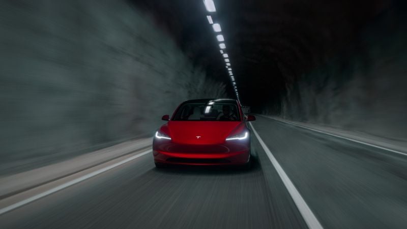 A red Tesla Model 3 shown from the front three-quarter angle driving through a dimly lit tunnel, with its distinctive LED headlights illuminated and creating a dramatic atmosphere.