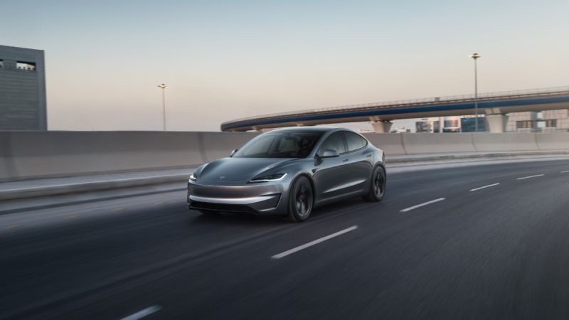 A gray electric car drives on a highway near an overpass at dusk.