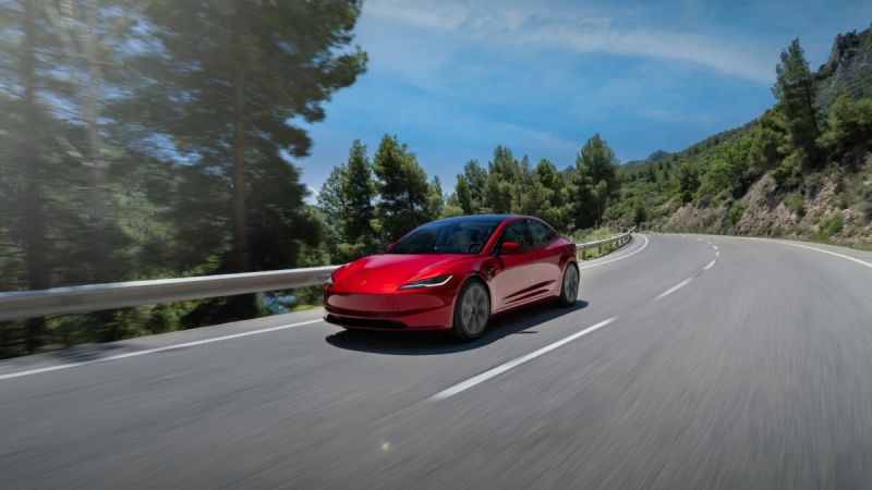 Candy red Tesla Model 3 driving on a winding mountain road, photographed from front three-quarter view, surrounded by pine trees and guardrails.