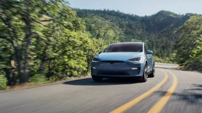 A light blue Tesla Model X SUV driving on a winding mountain road, captured from a front three-quarter angle with lush green forest scenery in the background.
