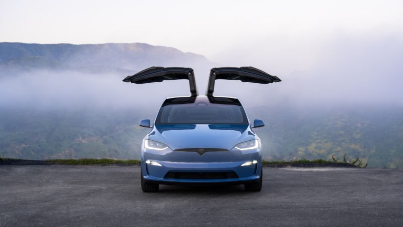 Blue Tesla Model X with signature falcon-wing doors open, front view, parked on mountain overlook with misty valley background.