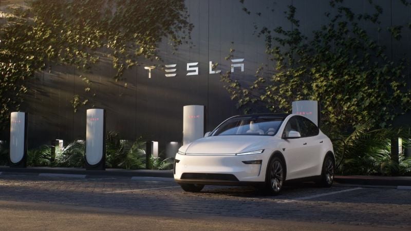 White Tesla Model Y parked at a Tesla Supercharger station with Tesla logo signage in the background.