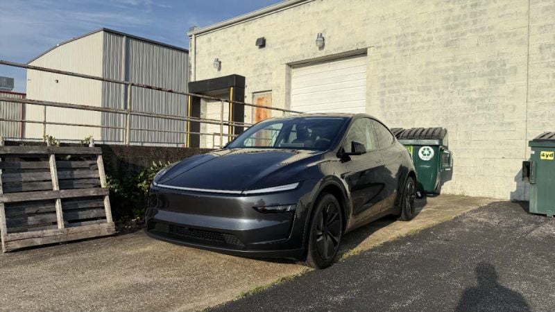 Black Tesla Model Y parked beside industrial building with recycling bins and wooden pallets