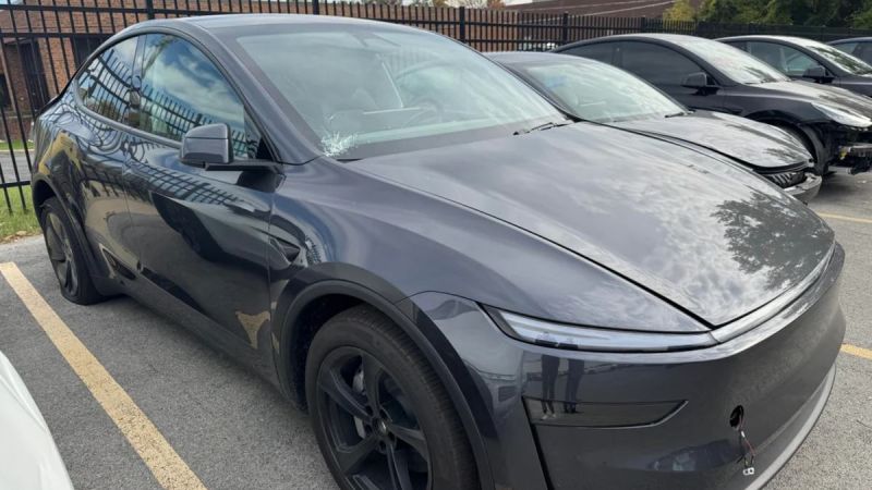 Gray Tesla Model Y Juniper, photographed at three-quarter front angle in parking lot, featuring sleek aerodynamic design, black wheels, and glass panoramic roof.