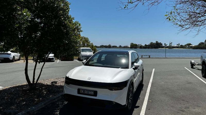 White car parked in a lot near a lake, surrounded by trees and other vehicles.