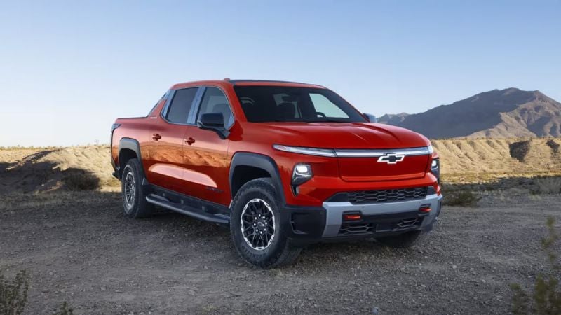 2026 Chevrolet Silverado EV electric pickup truck in red, front three-quarter view off-roading on desert terrain with rugged tires and bold grille design.