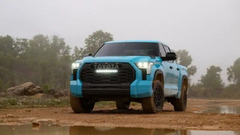 A head-on shot of a light blue 2025 Toyota Tundra TRD Pro with its distinctive LED light bar turned on, parked in a muddy, off-road environment.