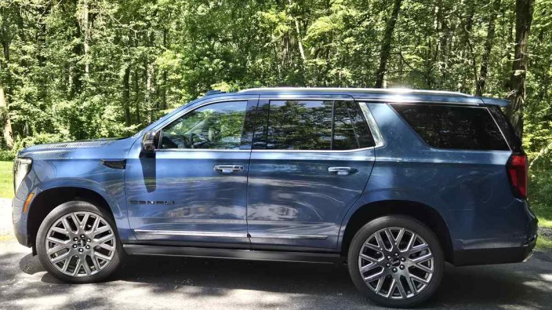 Blue 2025 GMC Yukon Denali SUV parked on asphalt with lush green forest background