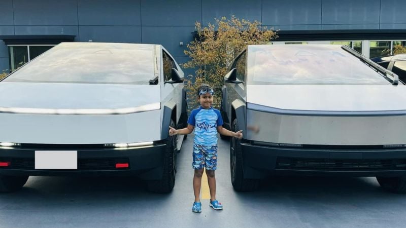 A child smiles between two Tesla Cybertrucks with metallic finishes. The scene is calm and set against a modern building with plants in the background.