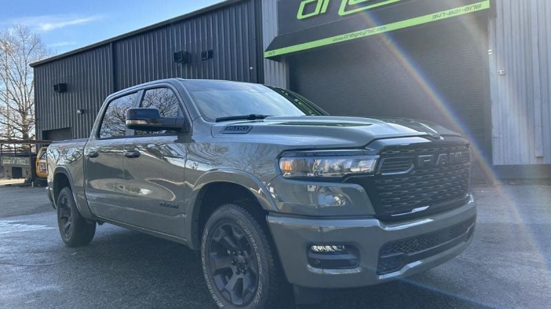 A gray 2026 Ram 1500 pickup truck shown from a front three-quarter angle, featuring black wheels and the aggressive RAM grille design, parked outside a building with a green sign.