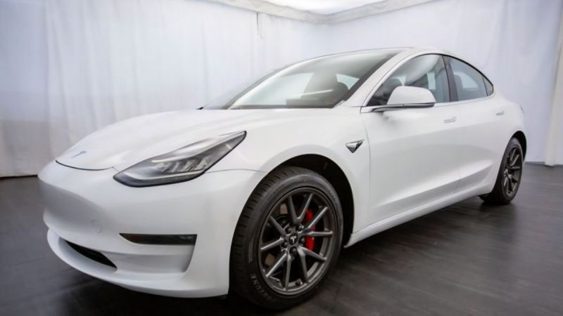A white Tesla Model 3 with a modern design is parked indoors on a showroom floor. It features aerodynamic curves, sharp headlights, and distinctive black wheels.