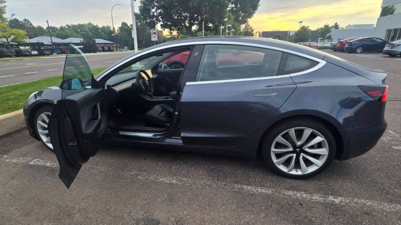 A dark grey Tesla Model 3 is parked with the driver’s door open under a cloudy sky. The setting is a quiet parking lot, evoking tranquility.