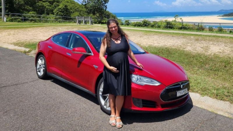 Anne Bishop in a black dress smiles while standing besides her red Tesla Model S on a scenic road. The background features greenery and a view of the ocean under a clear sky.