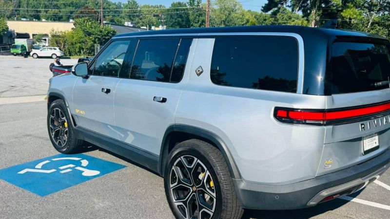 A gray Rivian R1S with tinted windows is parked in a handicap spot. The electric vehicle's sleek design and modern features are visible under a sunny sky.
