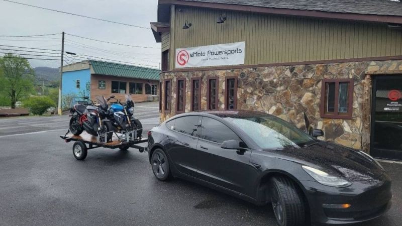 A black Tesla Model 3 is parked at a three-quarter front angle outside eMoto Powersports, with motorcycles on a trailer behind it.