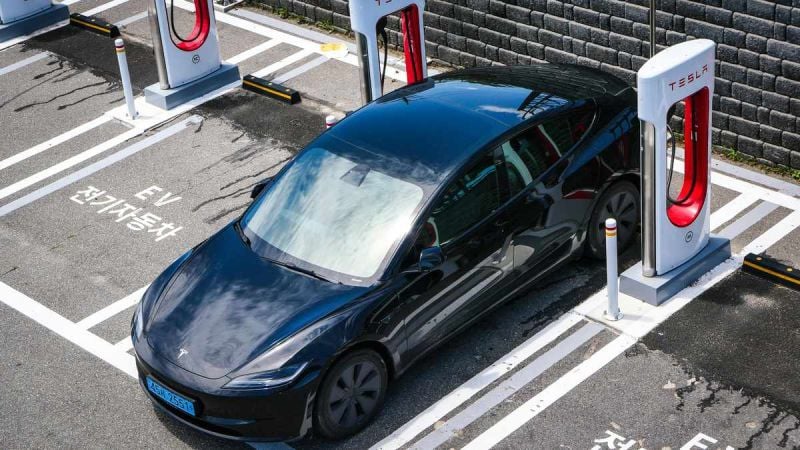 A black Tesla electric vehicle parked at a charging station, with wet pavement and marked EV parking spaces.