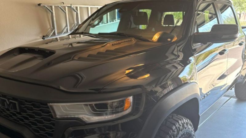 A shiny black 2025 Ram 1500 RHO is parked in a garage reflecting sunlight. Its aggressive design is accentuated by bold headlights and a textured hood.