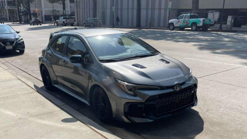 A dark gray Toyota GR Corolla is parked on a sunlit street. Its shiny exterior and sporty design convey a modern, dynamic feel. Nearby cars and city buildings form the backdrop.