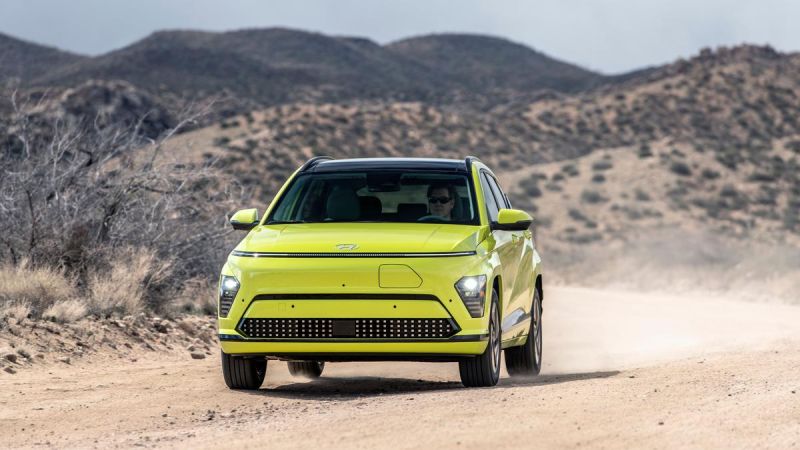 A vibrant green 2025 Hyundai Kona EV drives along a dusty road, surrounded by rugged hills and a cloudy sky.
