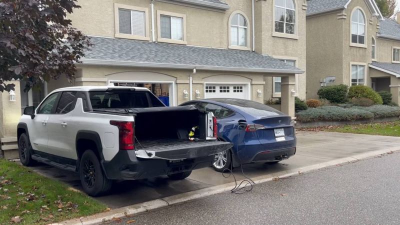 Blue Tesla Model Y being charged from white GMC pickup truck's bed outlet, rear three-quarter view, residential driveway setting.