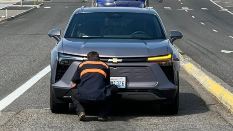 A person crouches in front of a silver Chevrolet Blazer EV on a road. They are focused on the front grille, with a road and another vehicle visible behind.