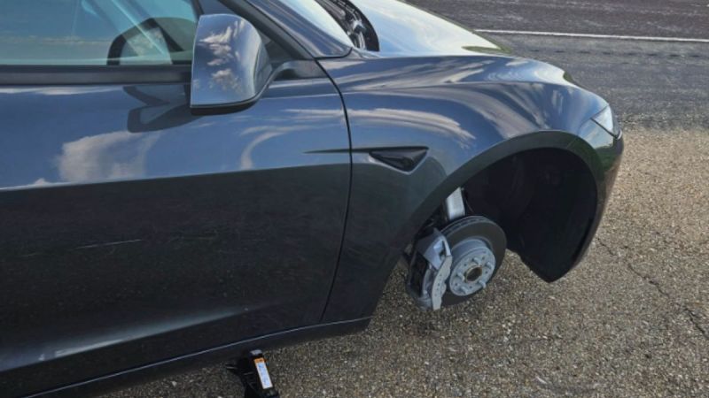 A black Tesla Model 3 parked roadside with front wheel missing, revealing the brake rotor and caliper. A car jack is also positioned beneath the vehicle.