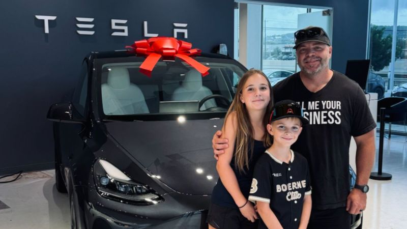 Clint's family stands in front of a black Tesla Model Y with a large red bow on top. They smile warmly in a Tesla showroom, conveying joy and excitement.