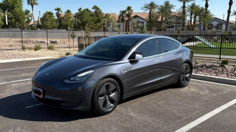 Sleek gray Tesla Model 3 parked in desert residential area, angled side view with palm trees and metal fence background