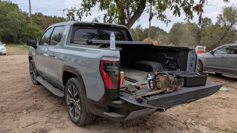 A gray GMC Sierra EV Denali parked on dirt with its tailgate down, carrying a chainsaw, toolbox, gloves, a drink can, and a water bottle on the bed
