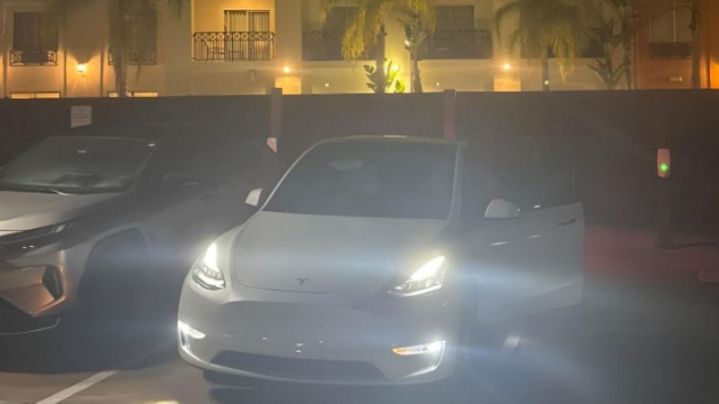 White Tesla Model Y with headlights on, parked in dimly lit lot with palm trees and building lights in background