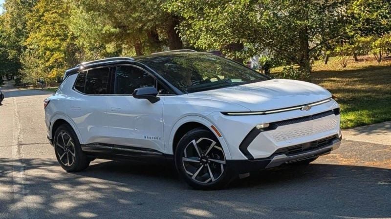 A white Chevrolet Equinox EV shown from a front three-quarter view, featuring black accents and distinctive two-tone wheels, parked in a shaded outdoor setting.