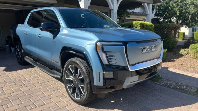 Light blue GMC Sierra Denali Ultimate pickup truck parked on brick driveway with landscaped background, showing bold front grille and premium styling