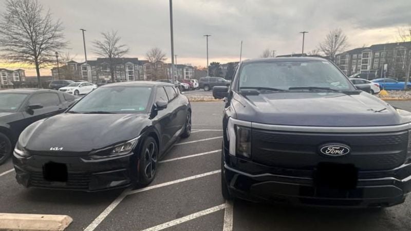 A black 2023 Kia EV6 and a dark blue Ford F-150 Lightning are parked side-by-side in a parking lot, both shown from their front three-quarter angles at dusk with apartment buildings visible in the background.