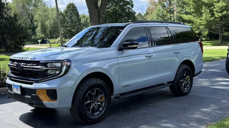 Elegant Grey Ford Expedition Tremor parked in a suburban driveway. The sun casts reflections on its sleek exterior. Green trees and a well-kept lawn surround the scene.